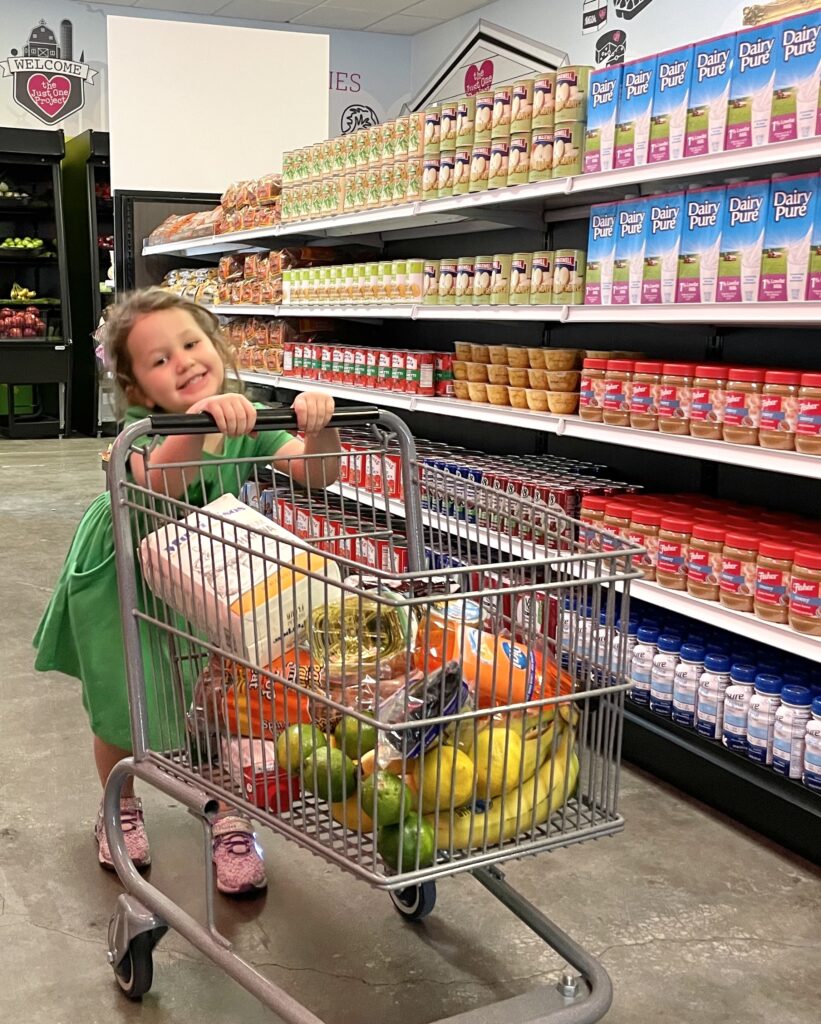 Young girl smiles as she pushes a cart through The Just One Project market.