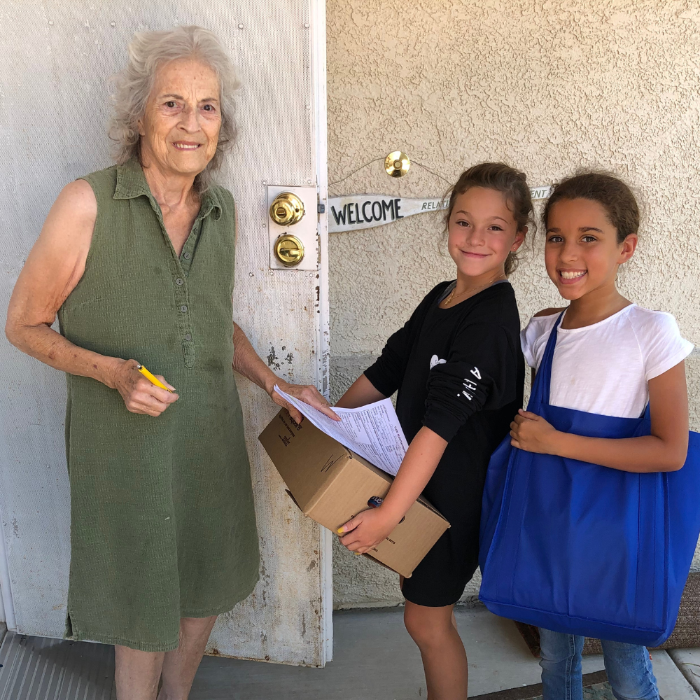 A woman and two girls standing in front of a door delivering groceries to a senior woman.
