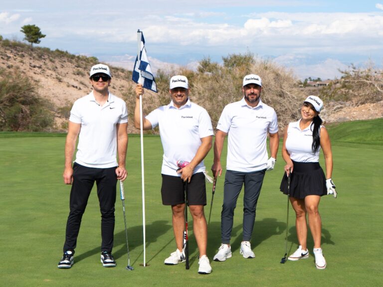 Four golfers stand together on a green golf course holding clubs and a flagstick, wearing matching white PacWest shirts and hats under a bright desert sky during The Just One Project’s 2025 Big Cup Golf Tournament.