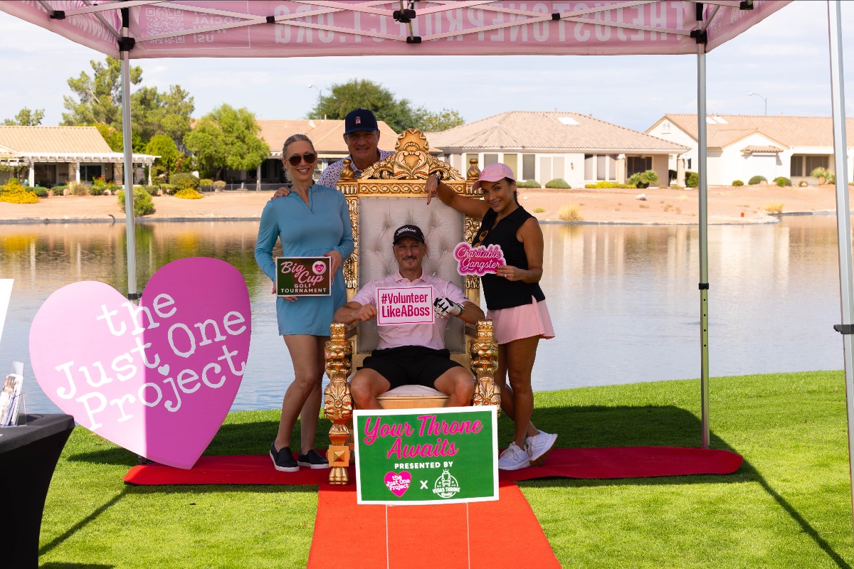A group of four golfers pose around a gold throne chair under The Just One Project’s pink event tent beside a lake, holding playful signs that read “Volunteer Like a Boss,” “Charitable Gangster,” and “Big Cup Golf Tournament.”