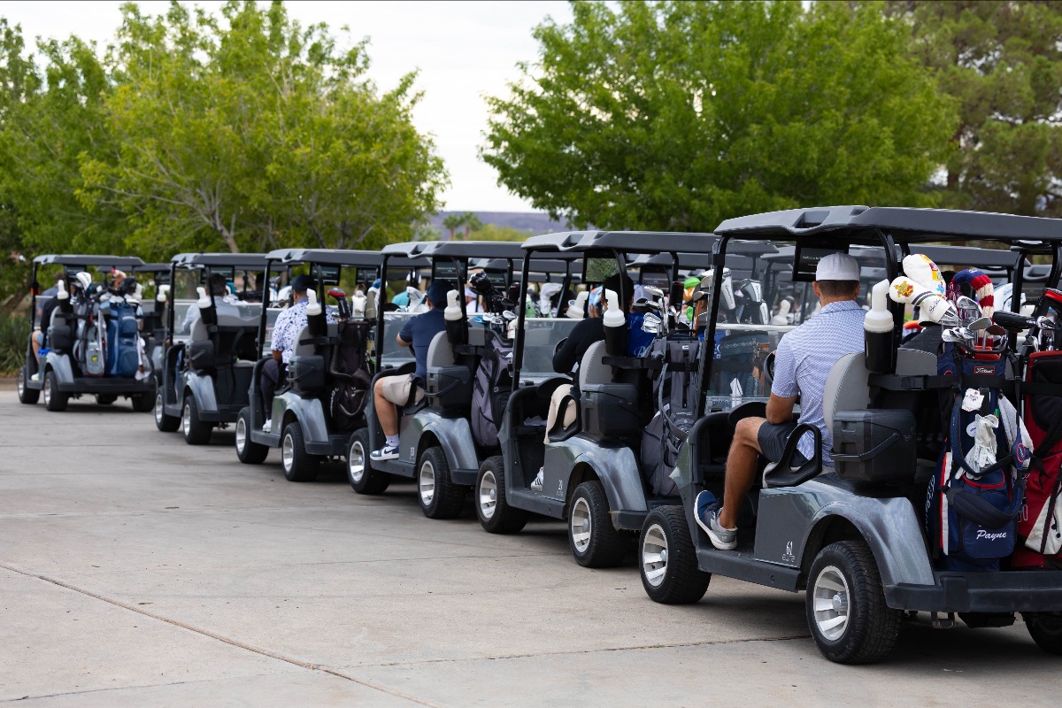 A long line of golf carts filled with players prepares for the shotgun start at The Just One Project’s 2025 Big Cup Golf Tournament, surrounded by lush green trees and desert scenery.