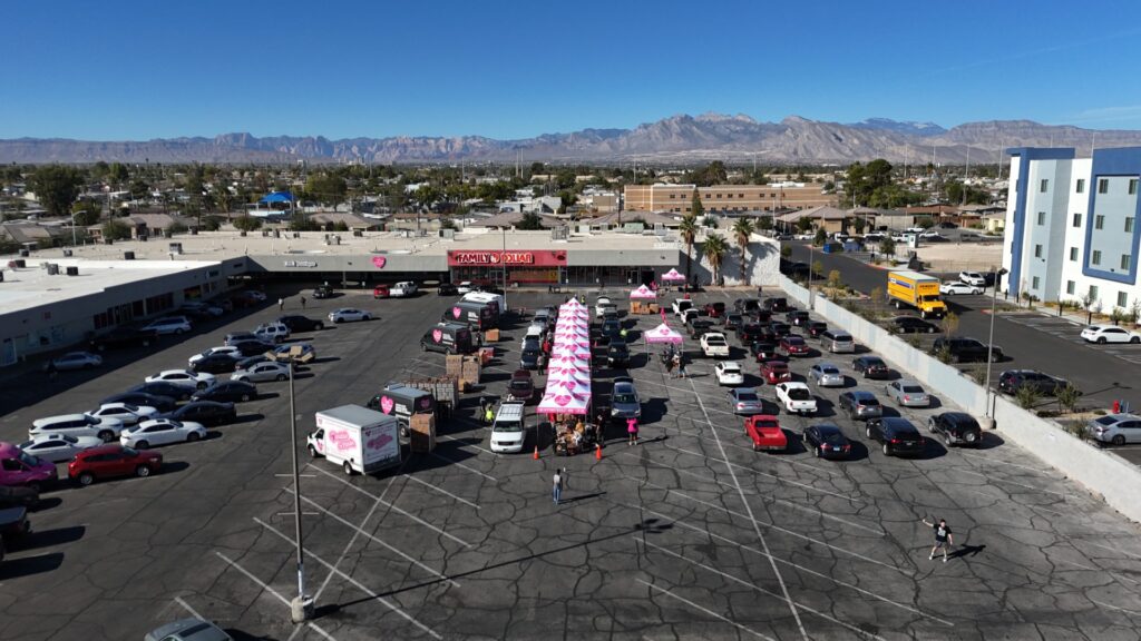 Aerial view of The Just One Project’s main distribution site with long lines of vehicles waiting for food assistance in a North Las Vegas parking lot; rows of pink TJOP-branded tents are surrounded by volunteers and parked cars as the city and mountains stretch in the background.