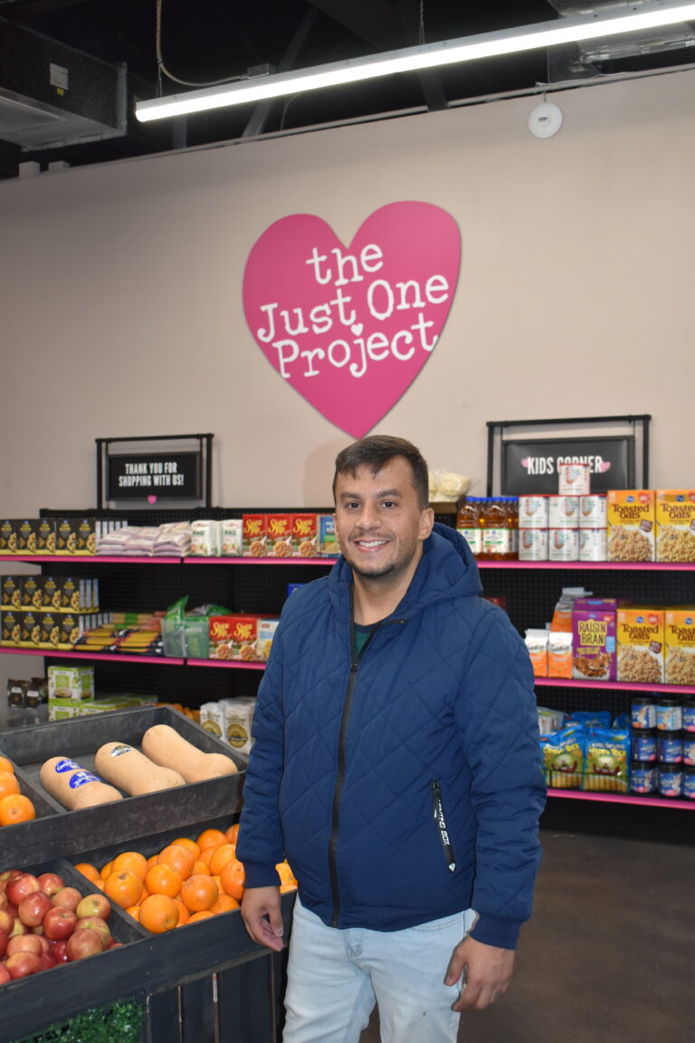 Romario smiling while standing inside The Just One Project’s community market, surrounded by fresh produce and pantry staples, after receiving housing stability support during his cancer treatment.